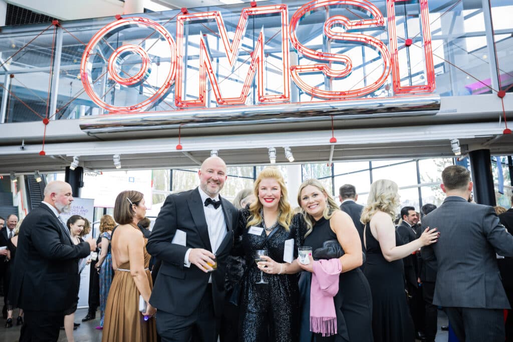Three people posing for a photo at a black-tie event under the neon OMSI sign in the museum's lobby